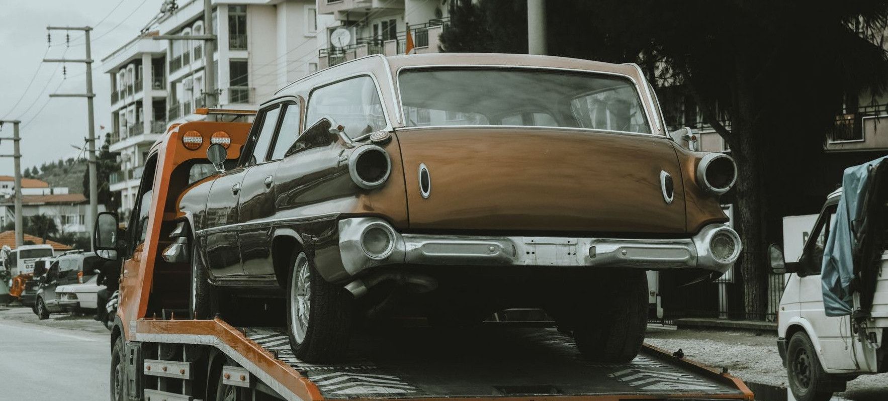 A vintage car loaded on a tow truck in a city street, surrounded by modern buildings.