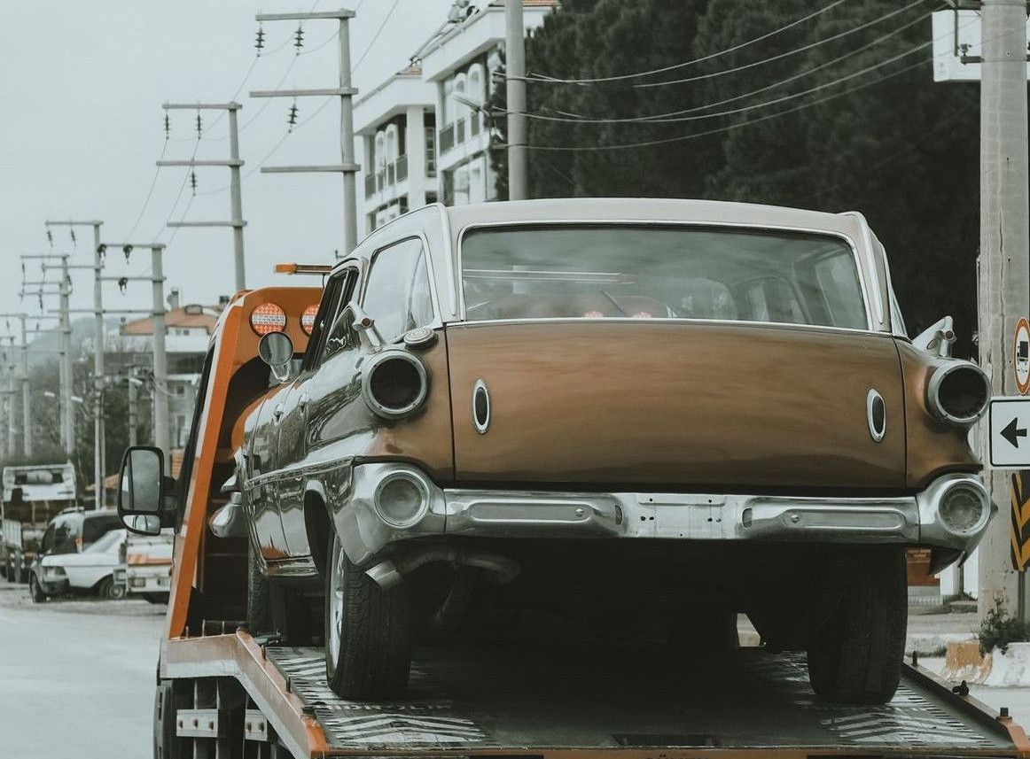 A vintage car is being towed through city streets on a tow truck, showcasing an urban environment.